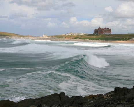 Large waves break at Fistral beach with a sandy shore and a red building on a clifftop beyond