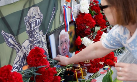 Woman lays flowers next to photo of Yevgeny Prigozhin and graphic painting of masked fighter