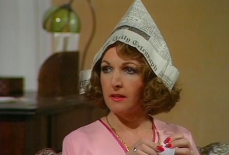 Head and shoulders of a woman with bright lipstick and eye make-up with permed hair and a pink top with a newspaper hat on her head