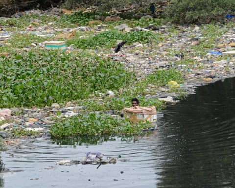 Um homem caminha por um rio cheio de lixo