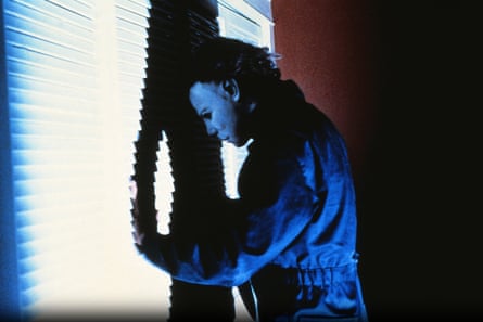 A man in a mask looks through Venetian blinds.