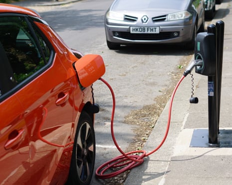 The corner of an orange car is shown; a red cable runs from behind an open flap in its rear wing to a roadside charging point mounted in the pavement.