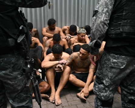 um grupo de jovens sem camisa sendo guardados por policiais armados