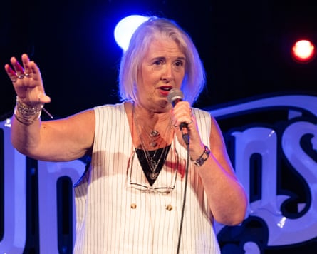 A woman in her 60s in a sleeveless top with a microphone, performing at a comedy show in Dublin
