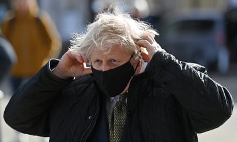 Boris Johnson outdoors in a coat, in the act of putting on a mask