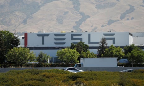 One-story beige white building with Tesla logo across it, with brown hills in the background.