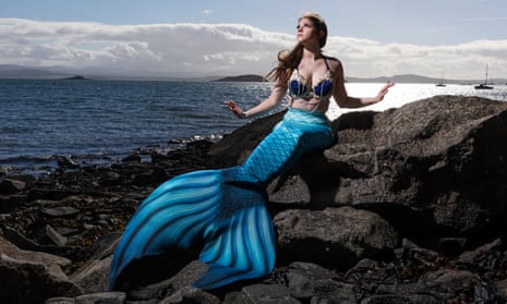 Ashleigh More, Scotland’s Moray Firth Mermaid, in her blue mermaid tail and bejewelled bikini top, sitting on rocks, looking ethereal towards the sky, her arms out like fins with the sea behind her