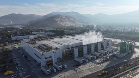 A large building surrounded by air vents which are sending plumes of steam into the air, with mountains in the background.