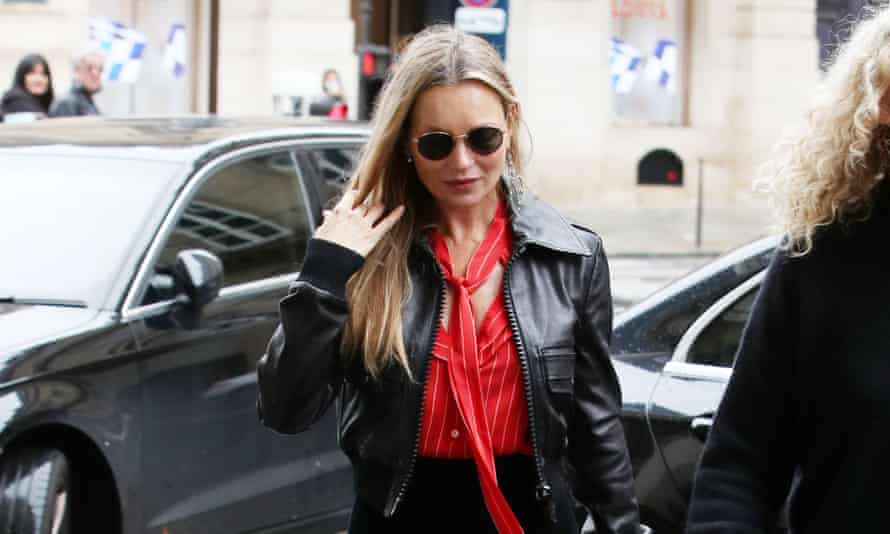 The model Kate Moss, wearing dark sunglasses, a black leather jacket, a red blouse and black trousers.