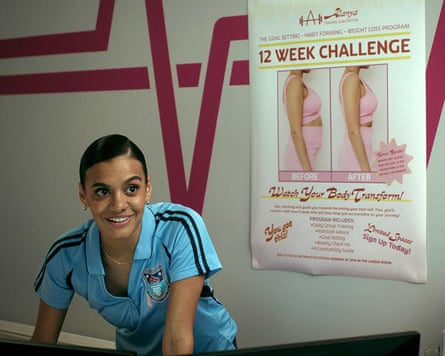 A woman in a sports shirt smiles in front of a poster that reads “12 week challenge” showing a photo of a woman in profile who is fat and another one of her slimmer