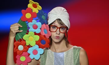 Models present Agatha Ruiz de la Prada's creation for the 2025 Spring/Summer Season in Madrid: a model wears large glasses, a white turban-style hat and striped shift dress, and holds a fan made up of big, colourful fabric flowers like a child's drawing