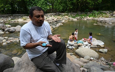 A man sits on a rock by a river, while children play in the water behind him.