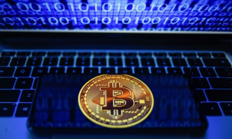 Photo illustration showing a bitcoin logo displayed on a smartphone with binary code on a laptop screen