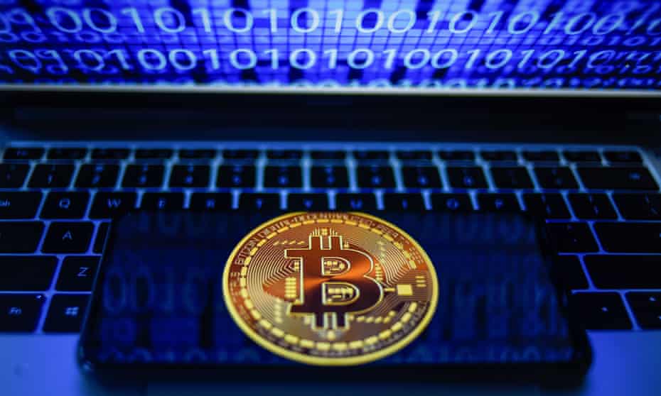 Photo illustration showing a bitcoin logo displayed on a smartphone with binary code on a laptop screen