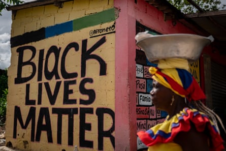 A Palenquera woman walks in front of graffiti related to the international social movement that fights against systemic racism and police brutality against Black people. On the right side, there are words in the Palenquera language that are used daily