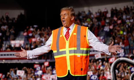 man wearing white white shirt, red tie and orange vest speaks and gestures wide with crowd of people looking on in the back