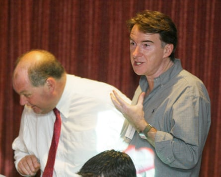 Roger Liddle in a white shirt and red tie bends down to speak to somebody out of shot while Peter Mandelson, in a grey shift gestures with his left hand.