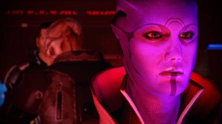In Mass Effect 2, a blue-skinned alien woman with facial markings looks forward, two figures blurred behind her