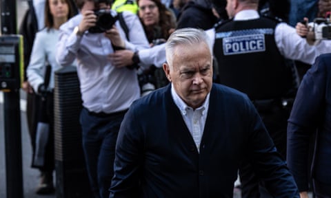 Huw Edwards walks towards court. Behind him a police officer is holding back a photographer