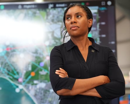 Badenoch stands looking serious, arms folded, before a screen showing a map with trajectories
