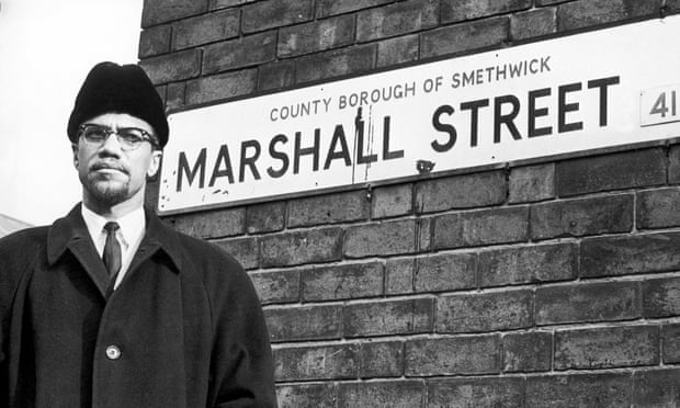 Malcolm X African-American Muslim minister and human rights activist poses beside the street sign for Marshall Street in Smethwick during a visit to the Midlands. The human rights activist posed beside the nameplate, walked 50 yards up the street, smiled at a “for sale” notice in the window of number 66 and returned to his car before being driven off. Malcolm X was assassinated nine days later on his return to the United States. 12th February 1965 (Photo by Birmingham Post and Mail Archive/Mirrorpix/Mirrorpix via Getty Images)