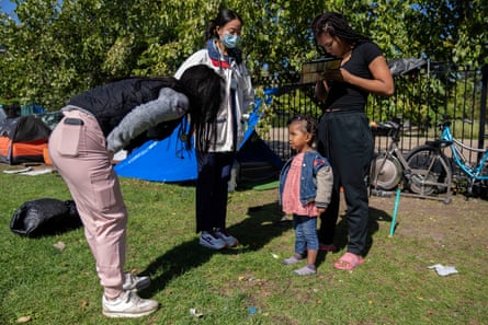 On a sunny lawn, three young women stand in a circle, with one leaning over to speak with a little girl with dark brown skin and black hair wearing a pink shirt and jeans jacket.