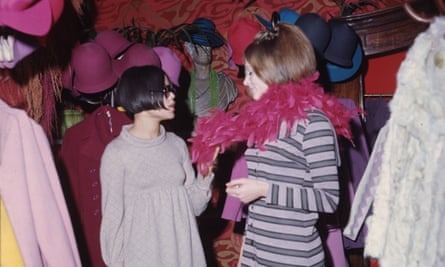 Two young women with bob-style hair. One is trying on a pink feather boa. Pink and blue hats are displayed on coat stands behind