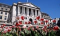 Red and white tulips are seen in front of the Bank of England building, seen from a low viewpoint against a bright, clear blue sky. The building is an imposing stone, classical-style structure with high arches and pillars.