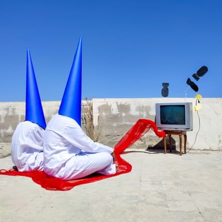 Two figures dressed in Sudanese traditional clothing and blue conical masks watch a TV with red fabric flowing out of it