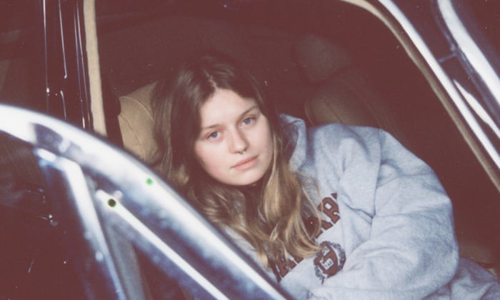 Photograph of girl in red, a white teenager girl with long dirty blond hair and a septum piercing. She's wearing a Harvard sweatshirt and lying down in the passenger seat of a car, looking at the camera.