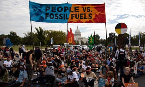 A crowd of protesters hold signs below a huge banner reading 'People vs fossil fuels'. The US Capitol building can be seen in the background.