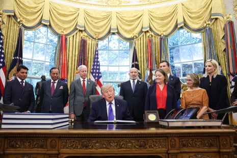 President Trump sits at his desk signing documents while officials stand behind him in the Oval Office