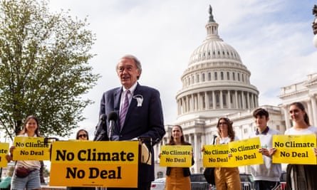 An older white man back by multiple young people in front of the white US Capitol dome, holding yellow signs that say, ‘No Climate, No Deal’.