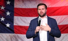 Young middle-aged white man with dark brown hair and full beard, wearing blazer, white shirt, no, tie, speaking into microphone he's holding in front of an American flag.