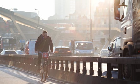 A man cycling along a cycle path on the edge of a busy road, with a flyover and buildings in the background