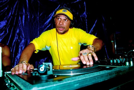 Goldie wearing a bright-yellow T-shirt and cap operating a mixing desk at an event