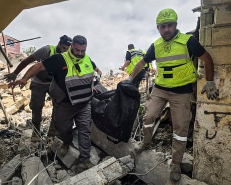 Three men carry a body bag through the rubble of a destroyed building.