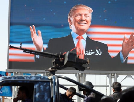 A truck with men in camouflage and a heavy machine gun mounted on the back pass a picture of a smiling Donald Trump on a big billboard