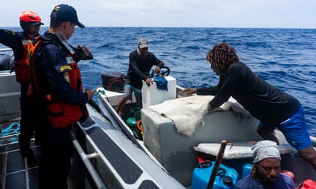Taken from a navy motorboat, two men on the left look into the smaller fishing boat alongside it on the right, where three men look to be showing what they have onboard.