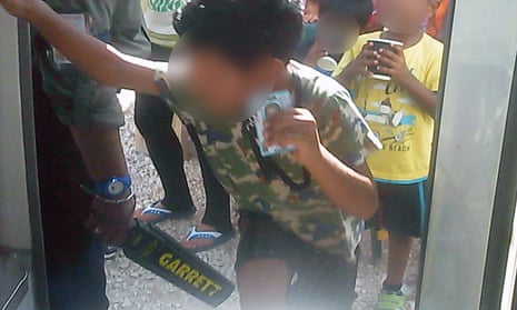 A young asylum seeker boy on Nauru holds up his ID card as a guard wands him. 