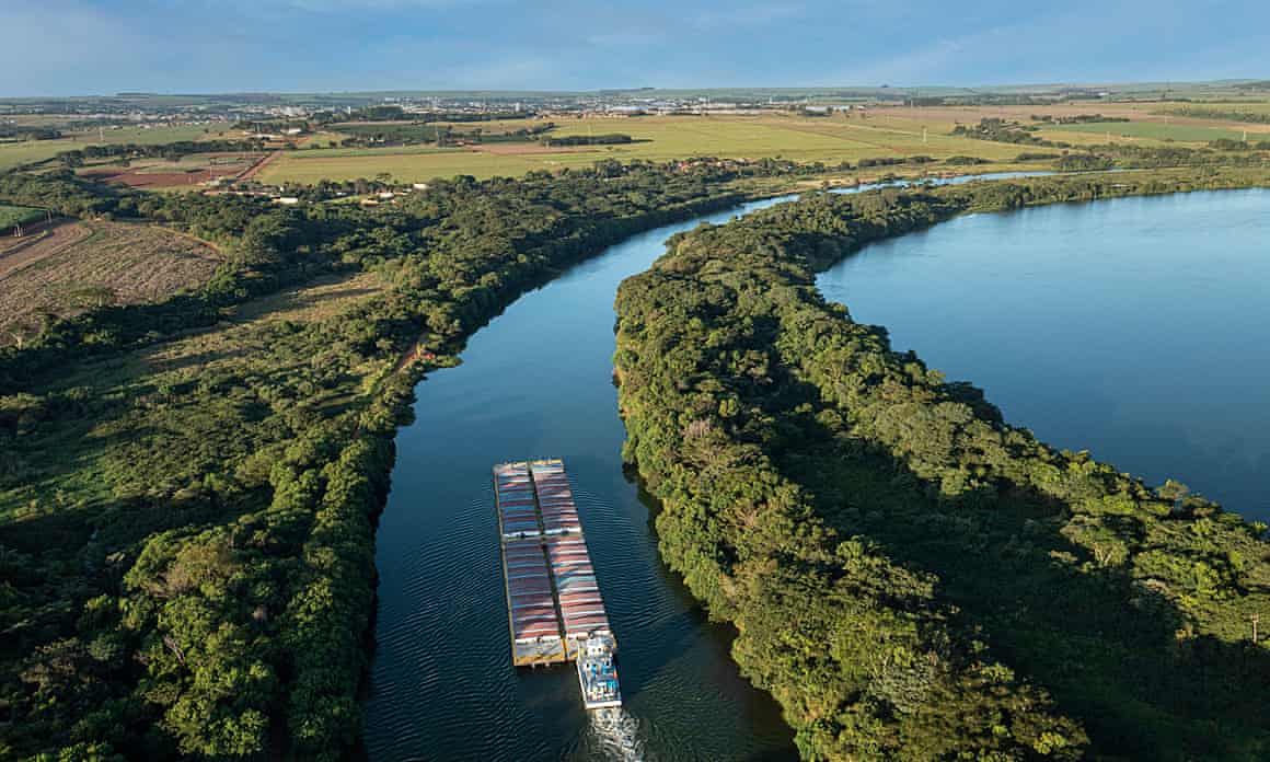 Shipping crates on a barge heading up a river surrounded by trees