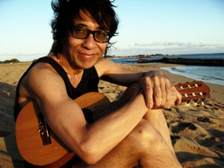 Makoto Kubota, former member of Les Rallizes Dénudés sitting on a beach holding a guitar.