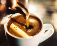 An espresso shot pours from the metal spout of an espresso machine into a small white cup of espresso