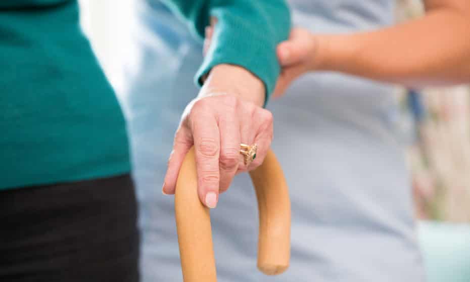 Senior Woman’s Hands On Walking Stick With Care Worker In Background