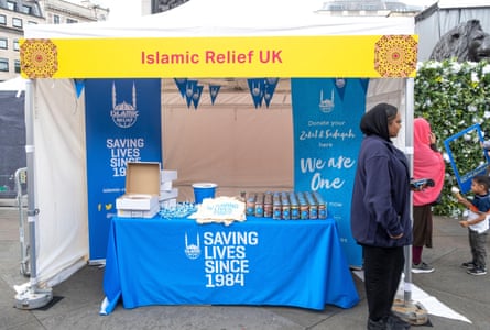 A woman holding a card machine stands at a stall holding cans of drink and blue lollipos and draped with blue banners reading ‘Saving lives since 1984’ and telling people that they can donate money.