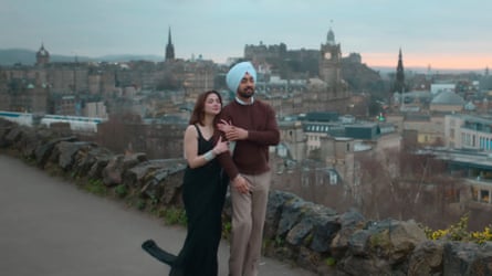 A man and a woman walk arm in arm as the sun sets over a city behind them.