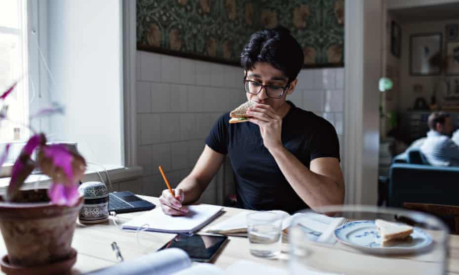 Young man of Asian background eating a sandwich while looking at mobile and writing on pad, while other people in soft focus sit on sofa.