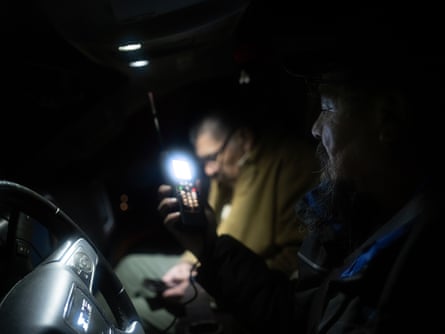 The faces of two men in a dark truck cabin are illuminated by a phone screen. John has a long beard.