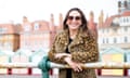 Bibi Lynch – journalist, writer and broadcaster –wearing a leopard print coat and sunglasses smiling leaning over a railing with pretty houses behind her