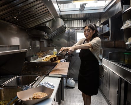 Awkwafina standing inside a food truck, holding food between two utensils and looking shocked